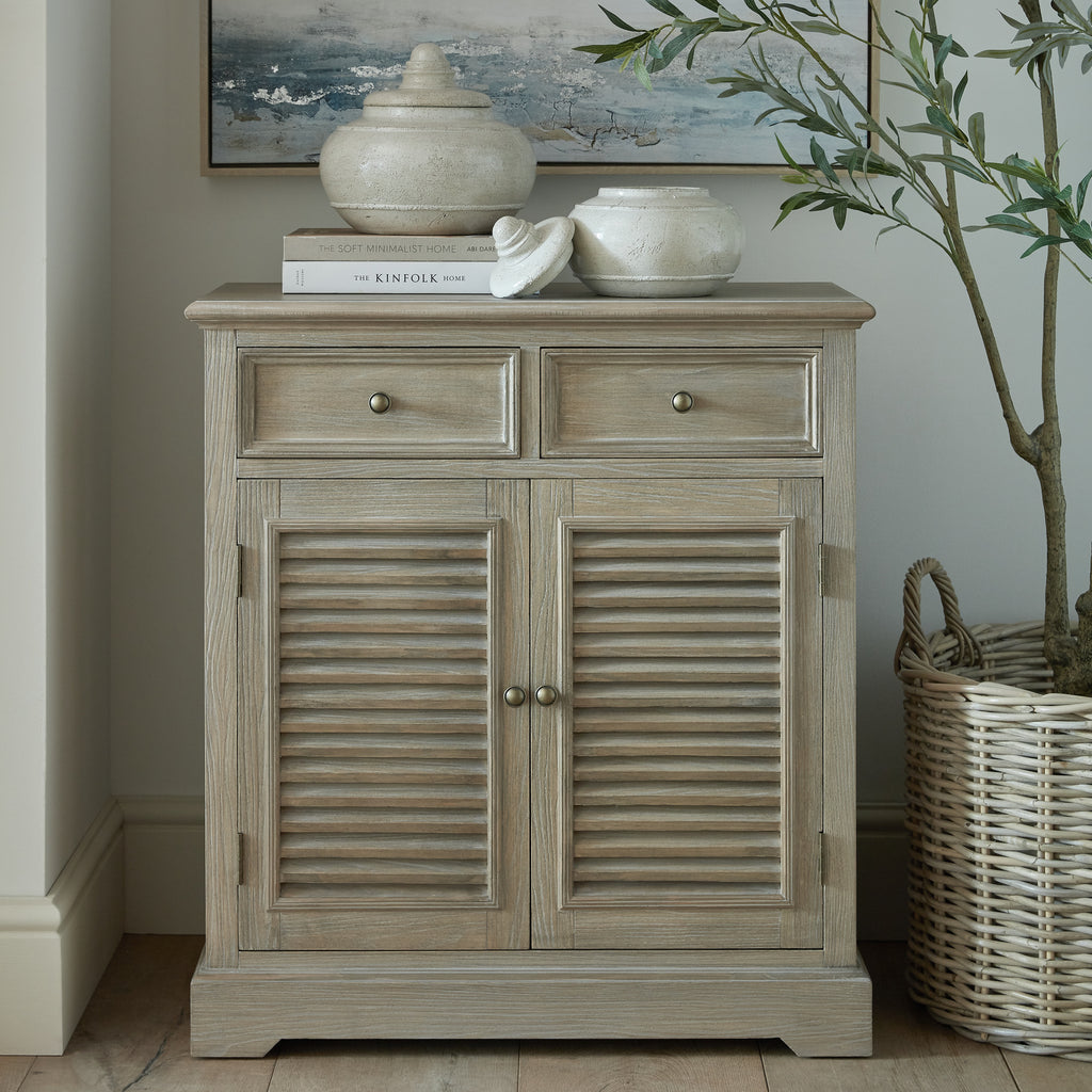 Louvred cabinet styled beneath a framed artwork, with pale decorative jars and books on top and a leafy tree in a woven basket beside it.
