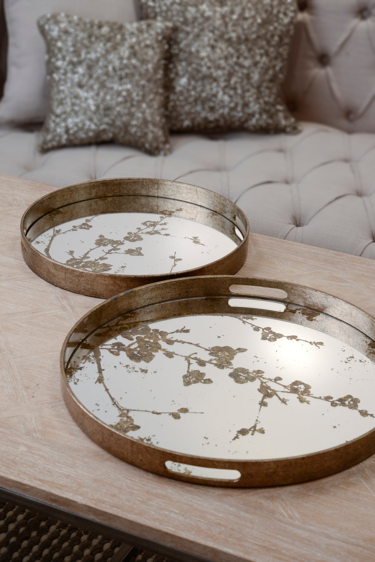 Two mirrored blossom trays on a pale wooden coffee table, with the larger tray in the foreground and a neutral buttoned sofa behind.