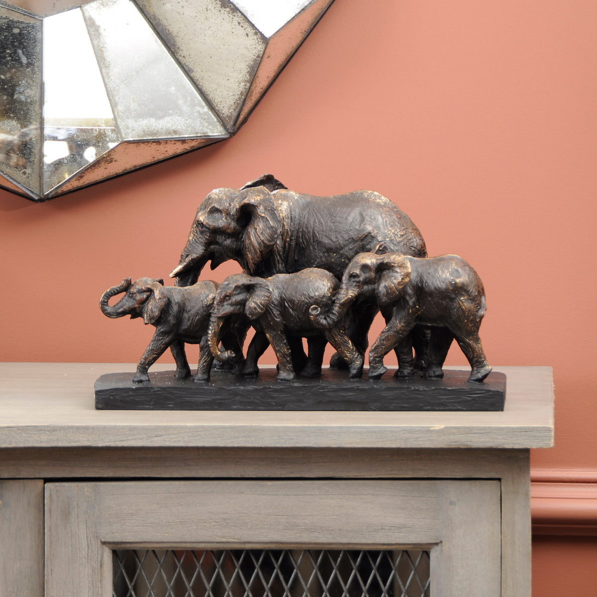 Matriarch elephant sculpture displayed on a pale wooden cabinet against a terracotta wall, beneath part of a geometric mirror.