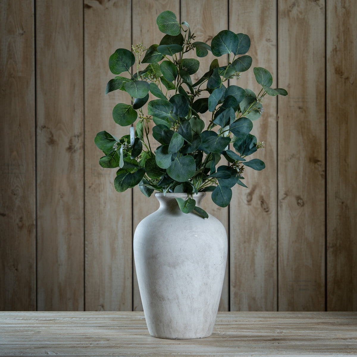 Grouped artificial eucalyptus foliage displayed in a pale vase on a wooden table against vertical wood panelling.