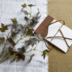 Mottled artificial foliage laid across white fabric beside an open brown notebook and pencil, showing olive and purple leaf tones.