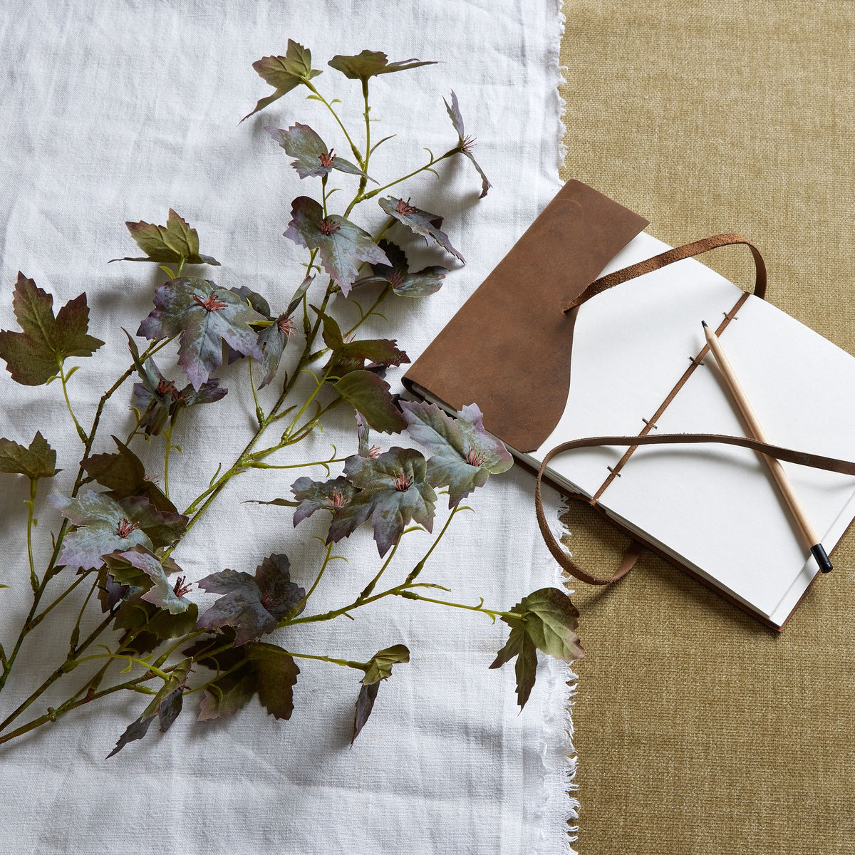 Mottled artificial foliage laid across white fabric beside an open brown notebook and pencil, showing olive and purple leaf tones.