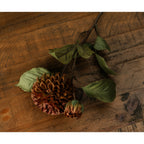Artificial flower stem laid diagonally on wood, with the rounded bloom and closed bud in the foreground and veined green leaves along the brown branches.