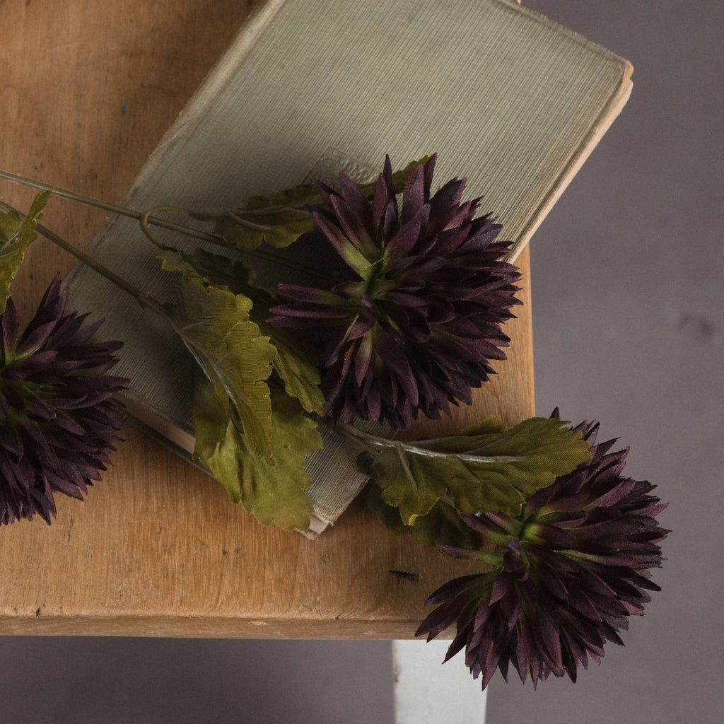 Close-up of dark purple artificial chrysanthemum blooms and lobed green leaves laid across a muted green book on a wooden table.