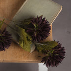 Close-up of dark purple artificial chrysanthemum blooms and lobed green leaves laid across a muted green book on a wooden table.
