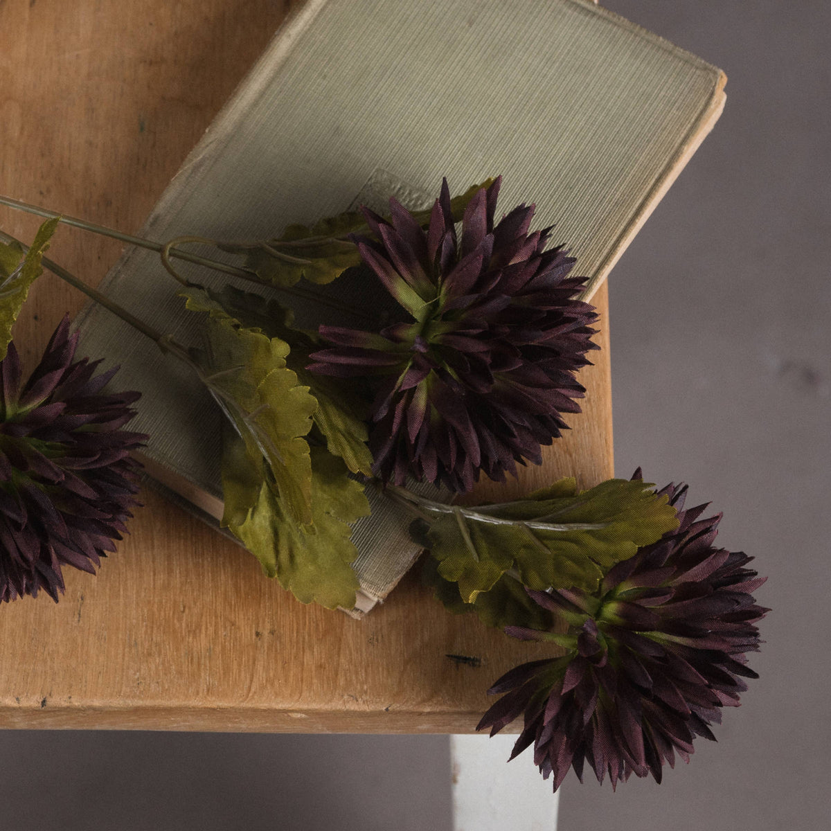 Close-up of dark purple artificial chrysanthemum blooms and lobed green leaves laid across a muted green book on a wooden table.