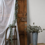 Pine garland hanging beside white draped fabric and a wooden door, trailing over a wooden sledge beside a bucket of decorative stems.