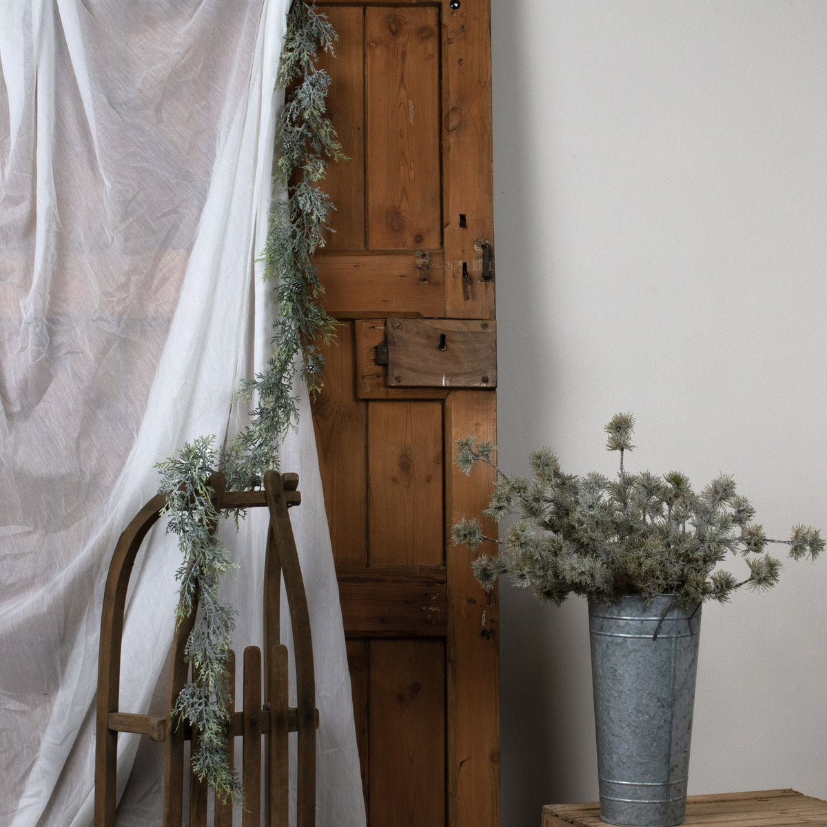 Pine garland hanging beside white draped fabric and a wooden door, trailing over a wooden sledge beside a bucket of decorative stems.
