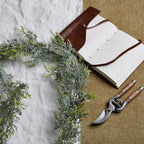 Frosted pine garland curved across white and tan fabric beside an open brown-covered notebook and pruning shears.