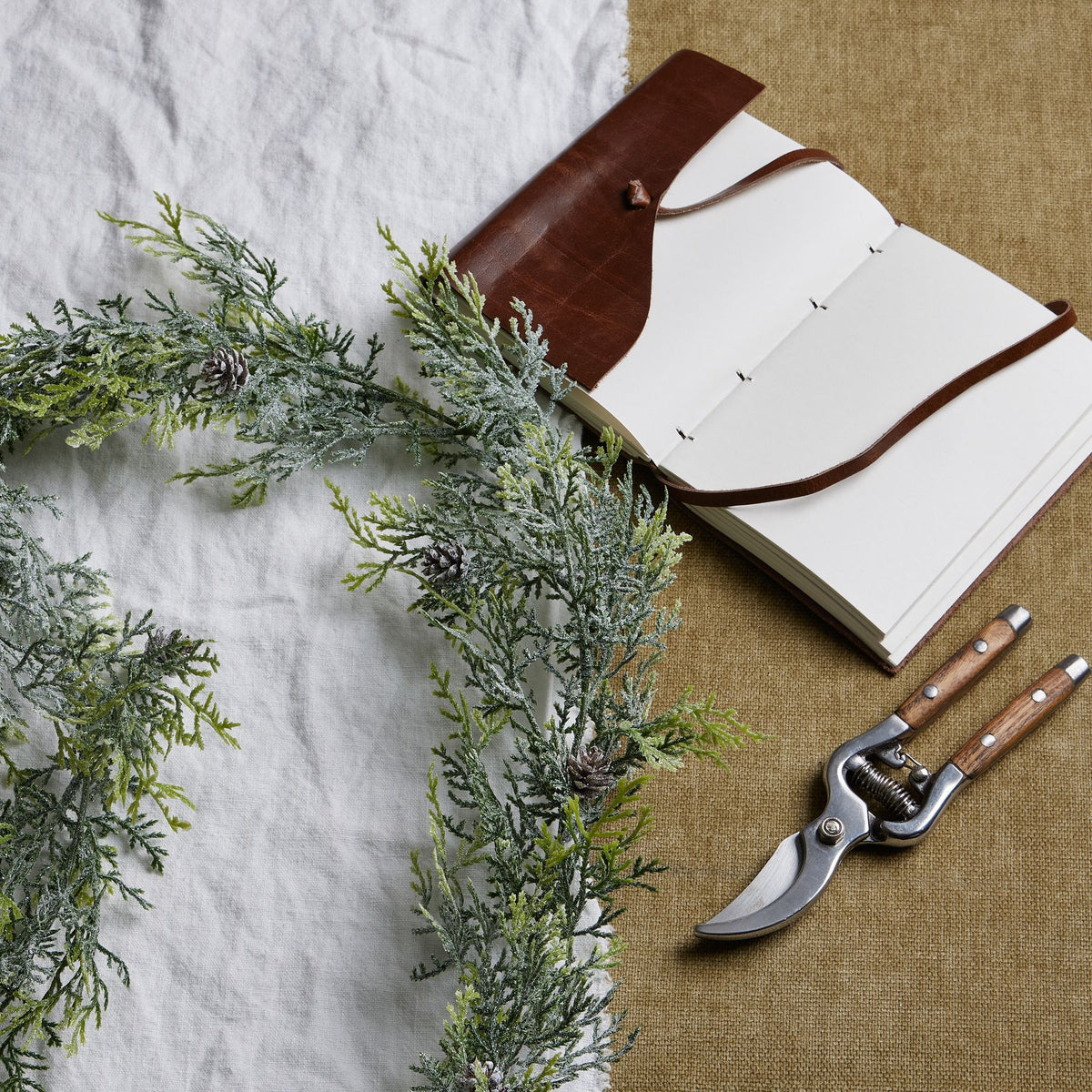 Frosted pine garland curved across white and tan fabric beside an open brown-covered notebook and pruning shears.