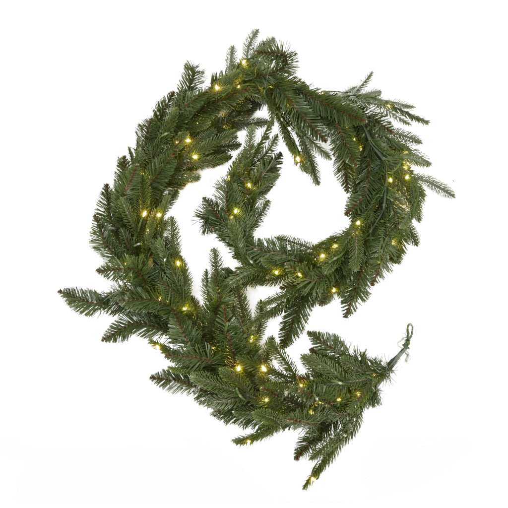 Green artificial garland arranged in a loose coil against white, with yellow lights visible through the needle foliage and an end loop at the lower right.