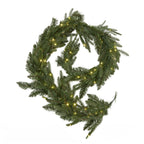 Green artificial garland arranged in a loose coil against white, with yellow lights visible through the needle foliage and an end loop at the lower right.