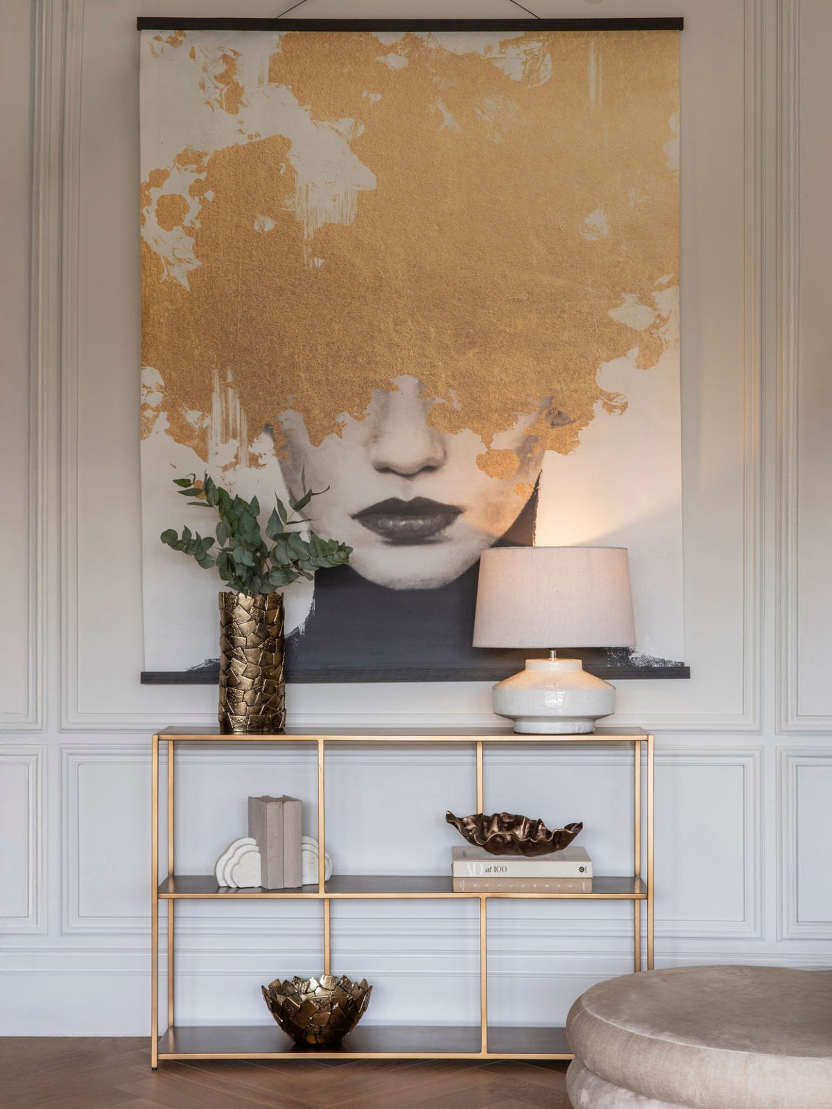 Portrait wall hanging with dark rails on a panelled wall above a gold-toned console, styled with a lamp, foliage, books and decorative bowls.