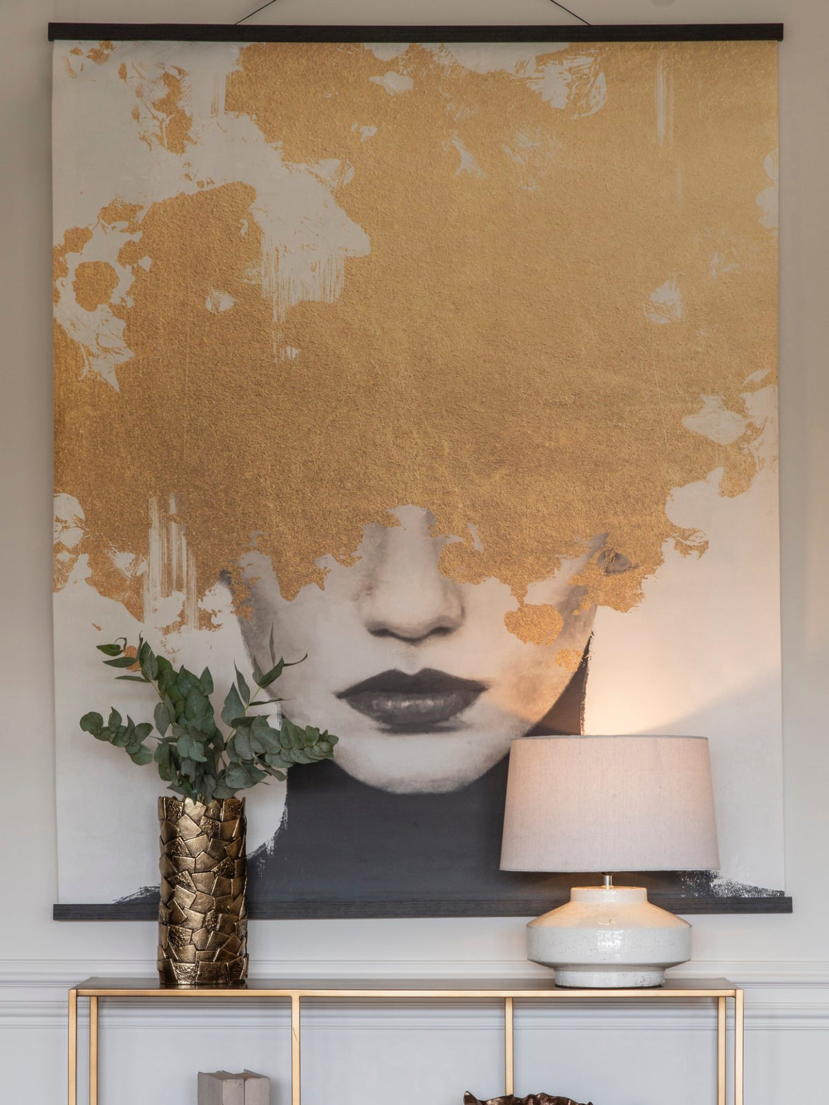 Close view of the portrait wall hanging with gold-coloured print obscuring the eyes, styled above a console with a leafy vase and table lamp.