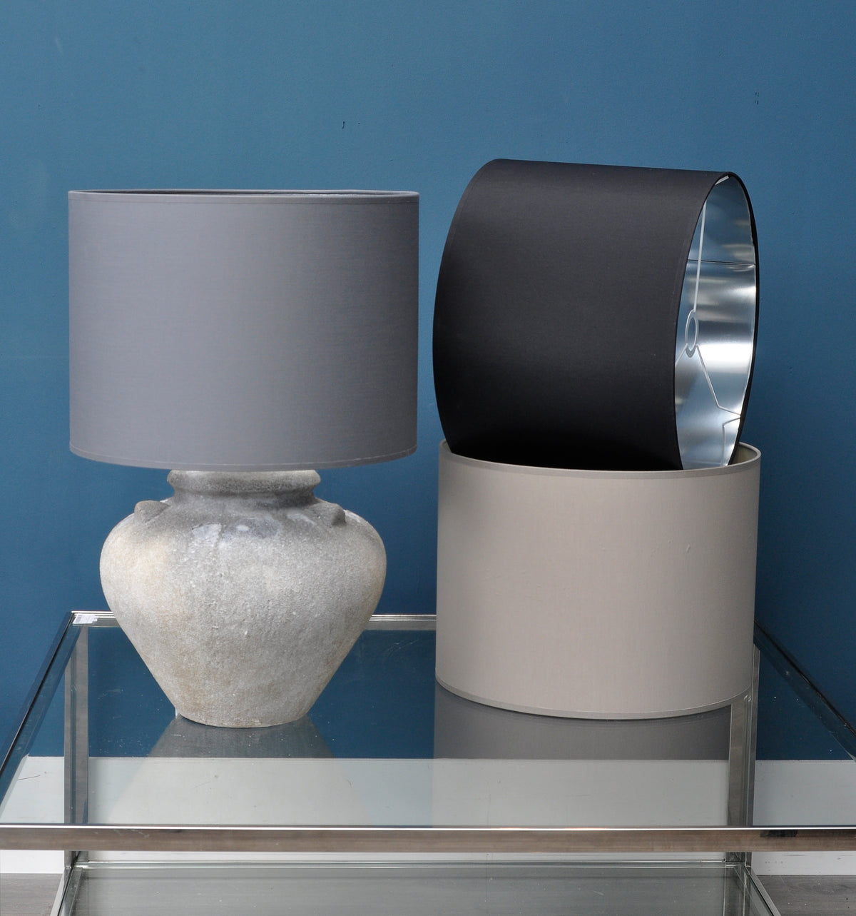 Black drum lampshade tilted inside a taupe shade, revealing its silver lining and white fitting, beside a grey-shaded lamp on a glass table.