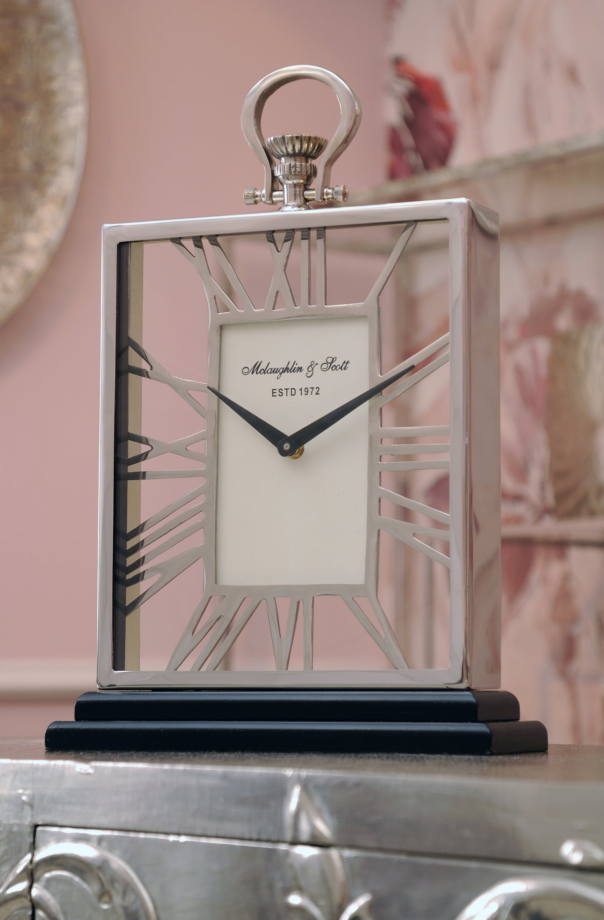 Noon Mantel Clock viewed at a slight angle on a reflective silver cabinet against a pink wall, with the case depth and open numeral design visible.