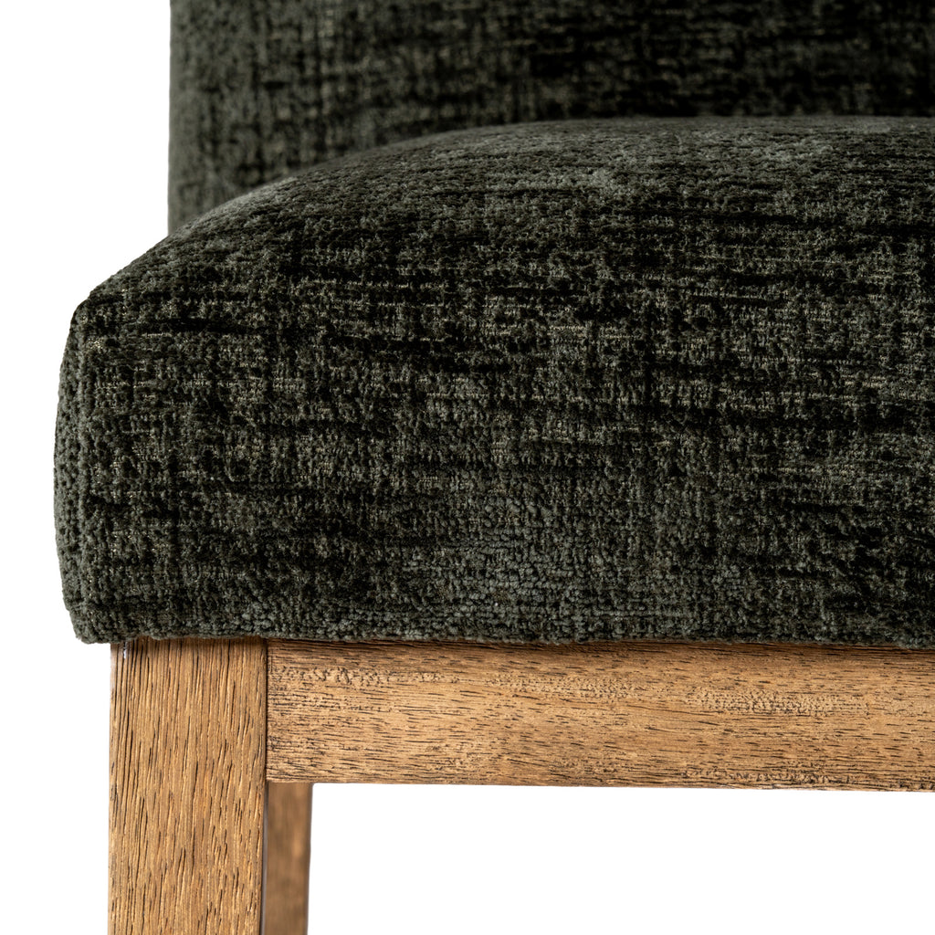 Close-up of the olive upholstered seat corner, showing its irregular dark texture above a warm brown rail and leg with visible grain.