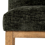 Close-up of the olive upholstered seat corner, showing its irregular dark texture above a warm brown rail and leg with visible grain.