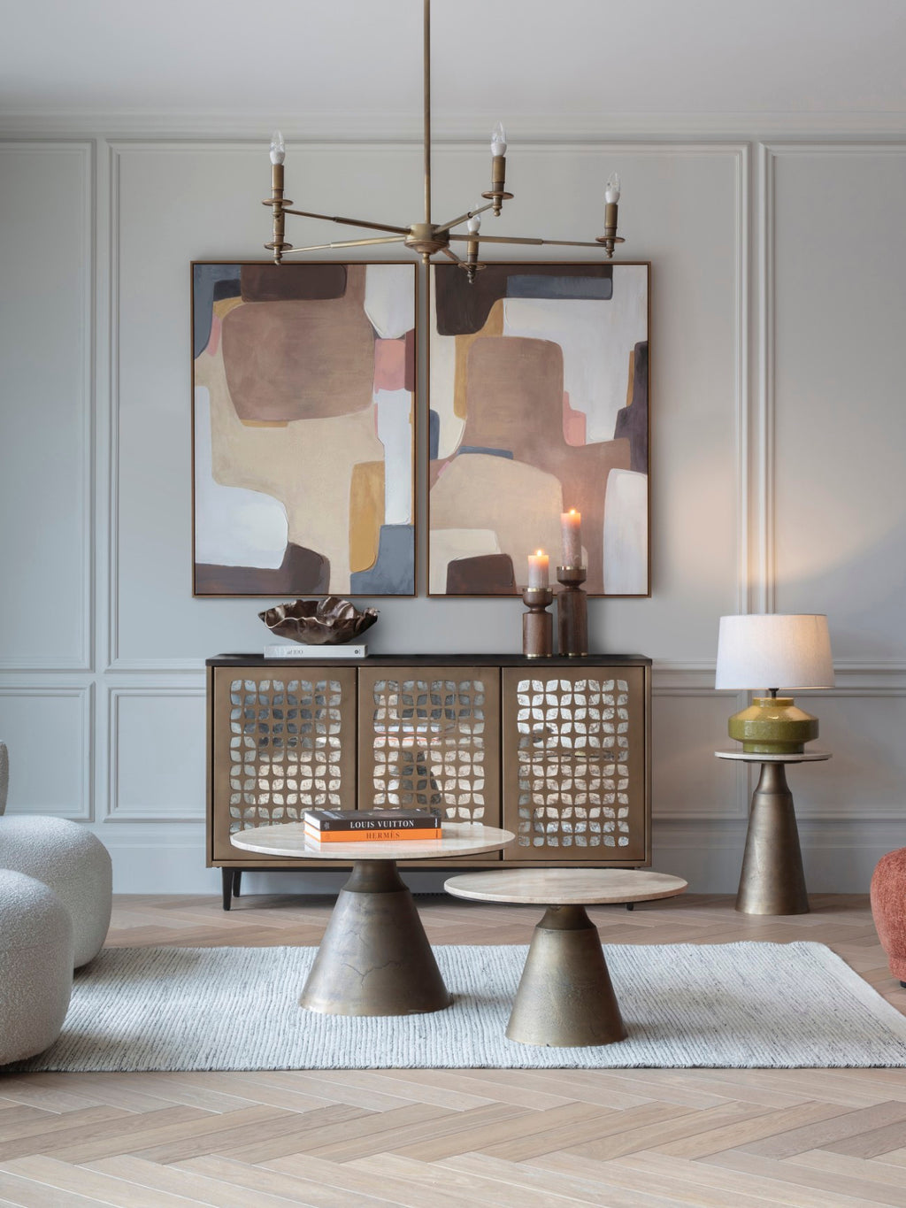 Open-arm brass chandelier in a living room with pale panelled walls, two abstract paintings, a mirrored cabinet and round coffee tables.