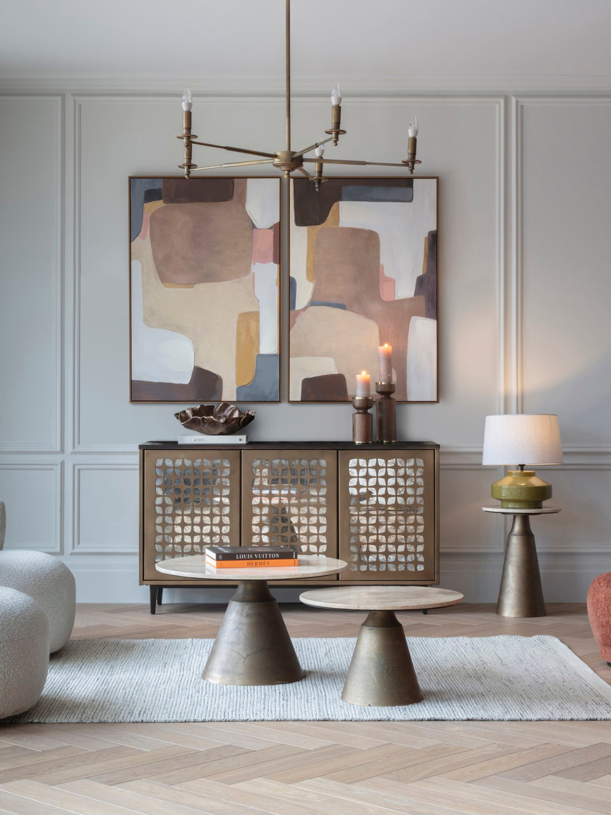 Open-arm brass chandelier in a living room with pale panelled walls, two abstract paintings, a mirrored cabinet and round coffee tables.