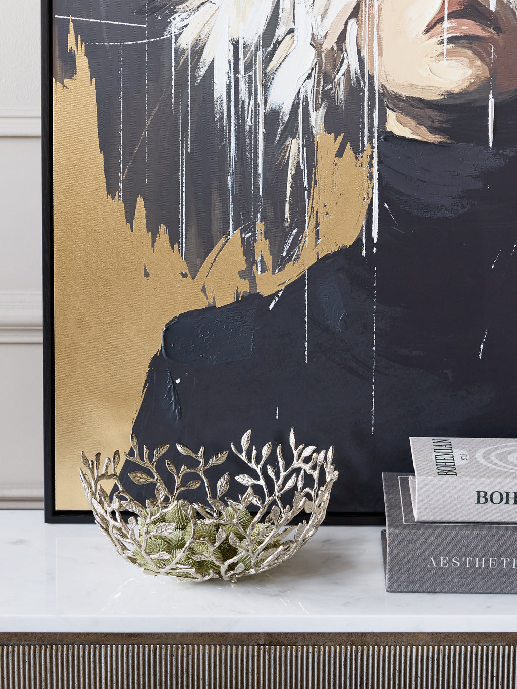 Open branch bowl holding green decorative foliage on a pale console beside books, with black and gold artwork behind.