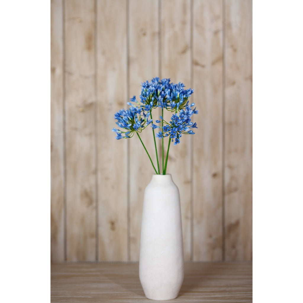 An arrangement of blue artificial flowers in a tall white vase on a wooden surface, with pale wooden panelling behind.