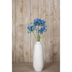An arrangement of blue artificial flowers in a tall white vase on a wooden surface, with pale wooden panelling behind.