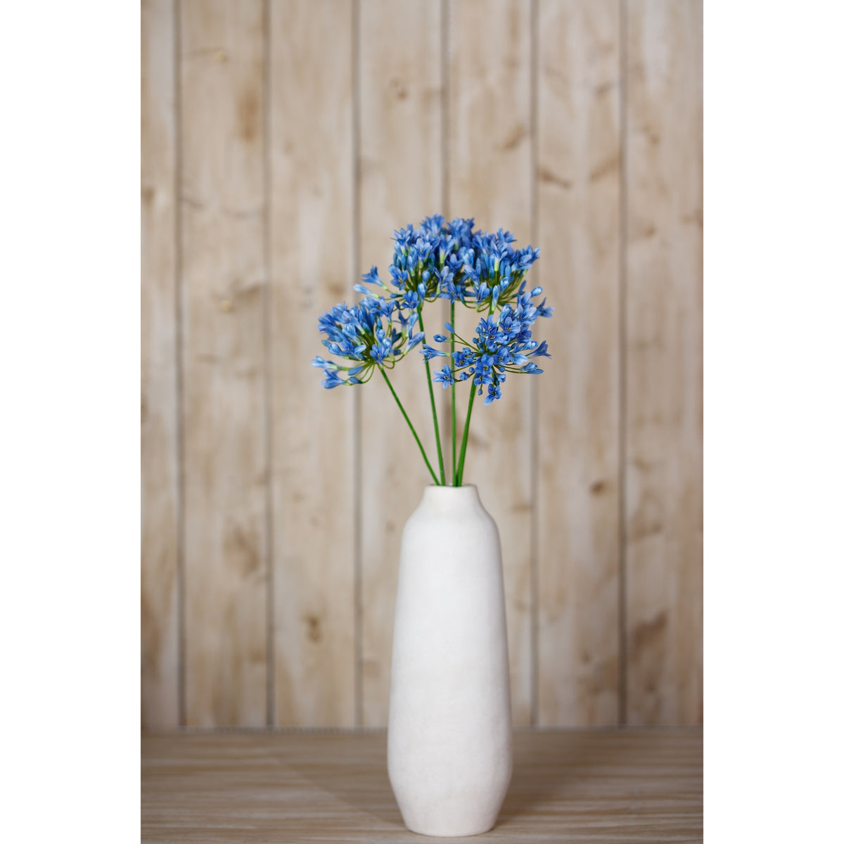 An arrangement of blue artificial flowers in a tall white vase on a wooden surface, with pale wooden panelling behind.