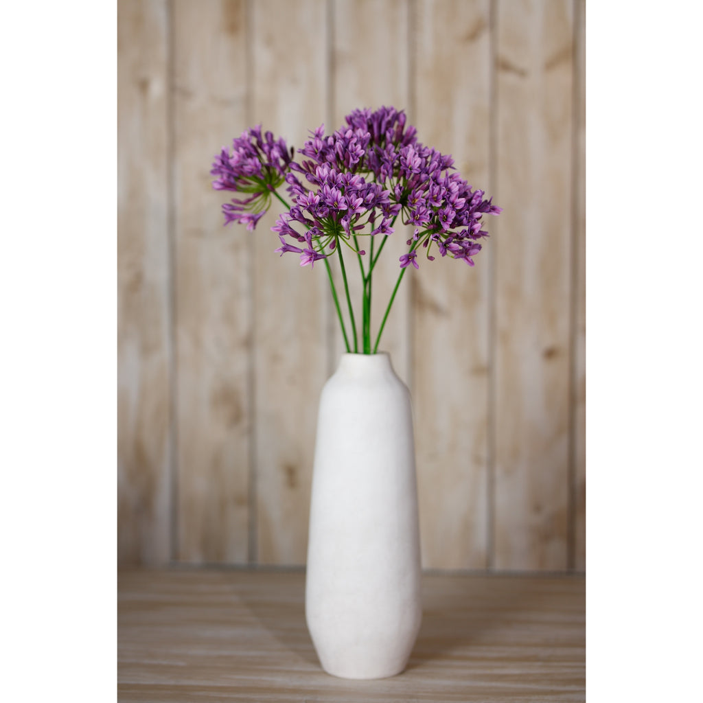 Purple-lilac artificial flowers arranged in a tall white vase on a wooden surface in front of pale vertical panelling.