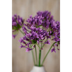 Detail of purple-lilac artificial florets with small yellow centres, with more flower heads blurred behind and the white vase partly visible below.