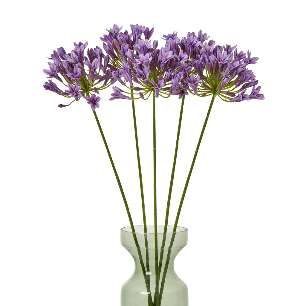 Several purple-lilac artificial flower stems spread across a translucent green vase against a white background.