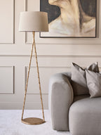 Gold triangle floor lamp beside a grey sofa against a pale panelled wall with a large abstract painting.