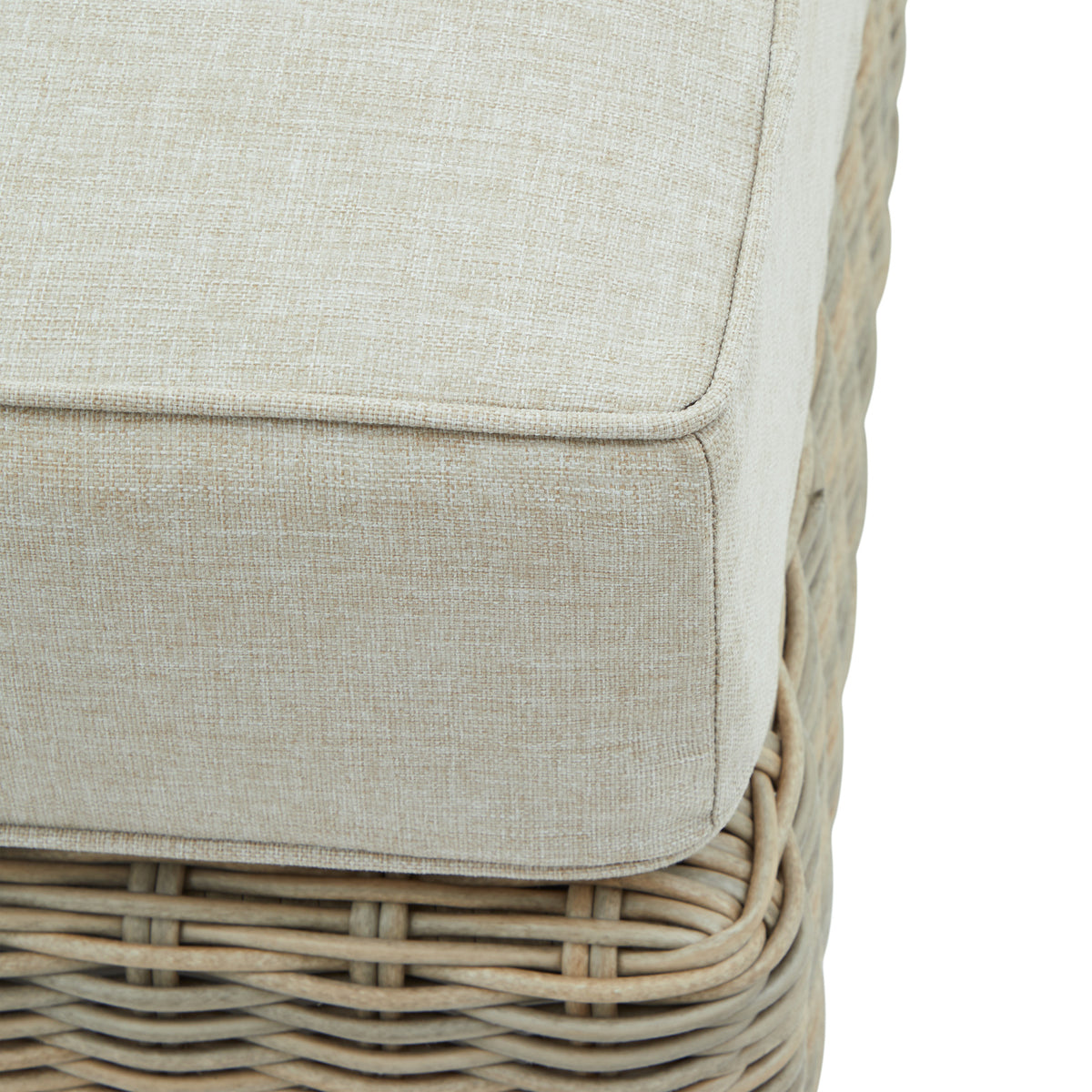 Close-up of the pale cushion's textured fabric and piped corner above the curved weave of the footstool base.
