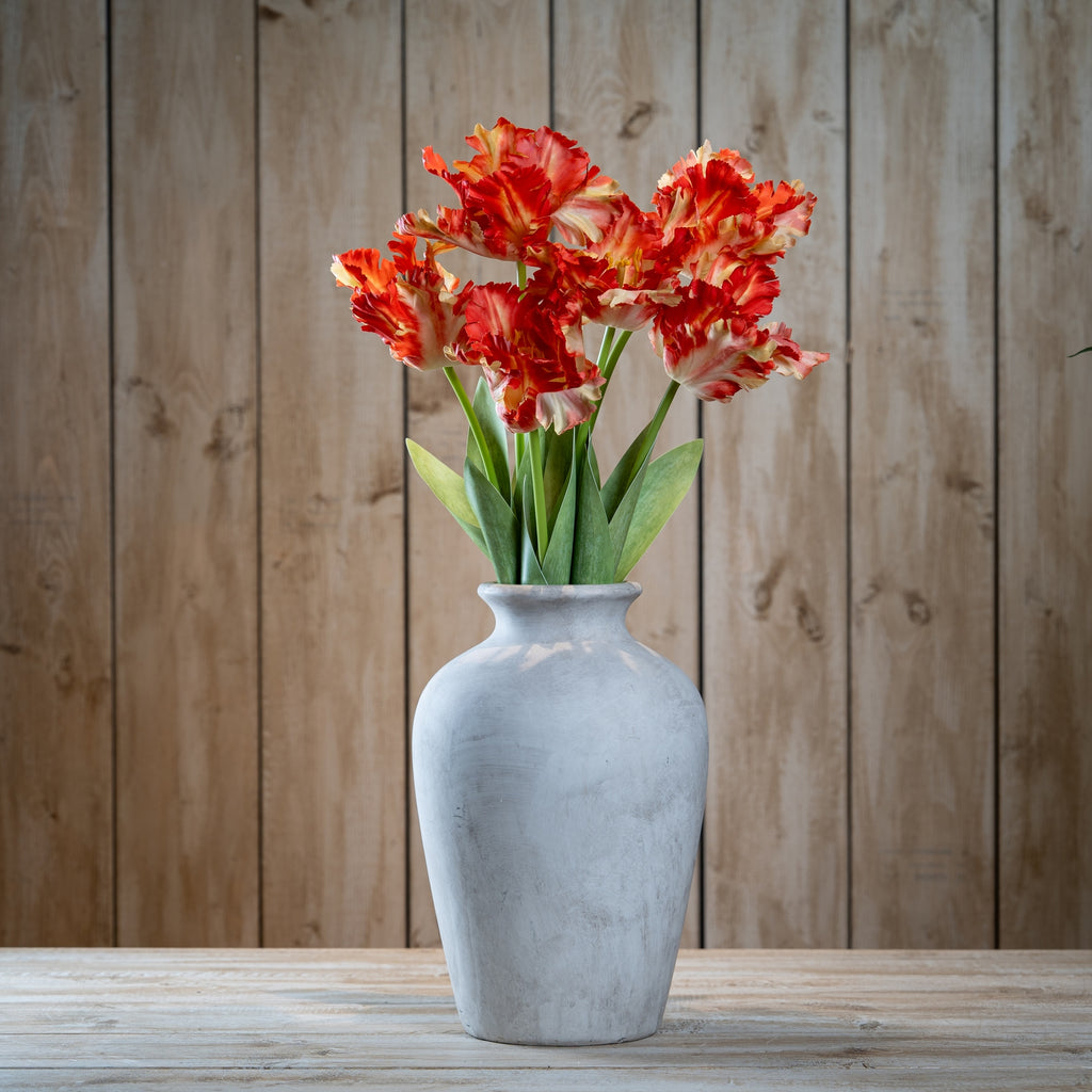 Several orange-red and cream artificial flower stems arranged in a pale grey vase on a wooden table against vertical timber boards.