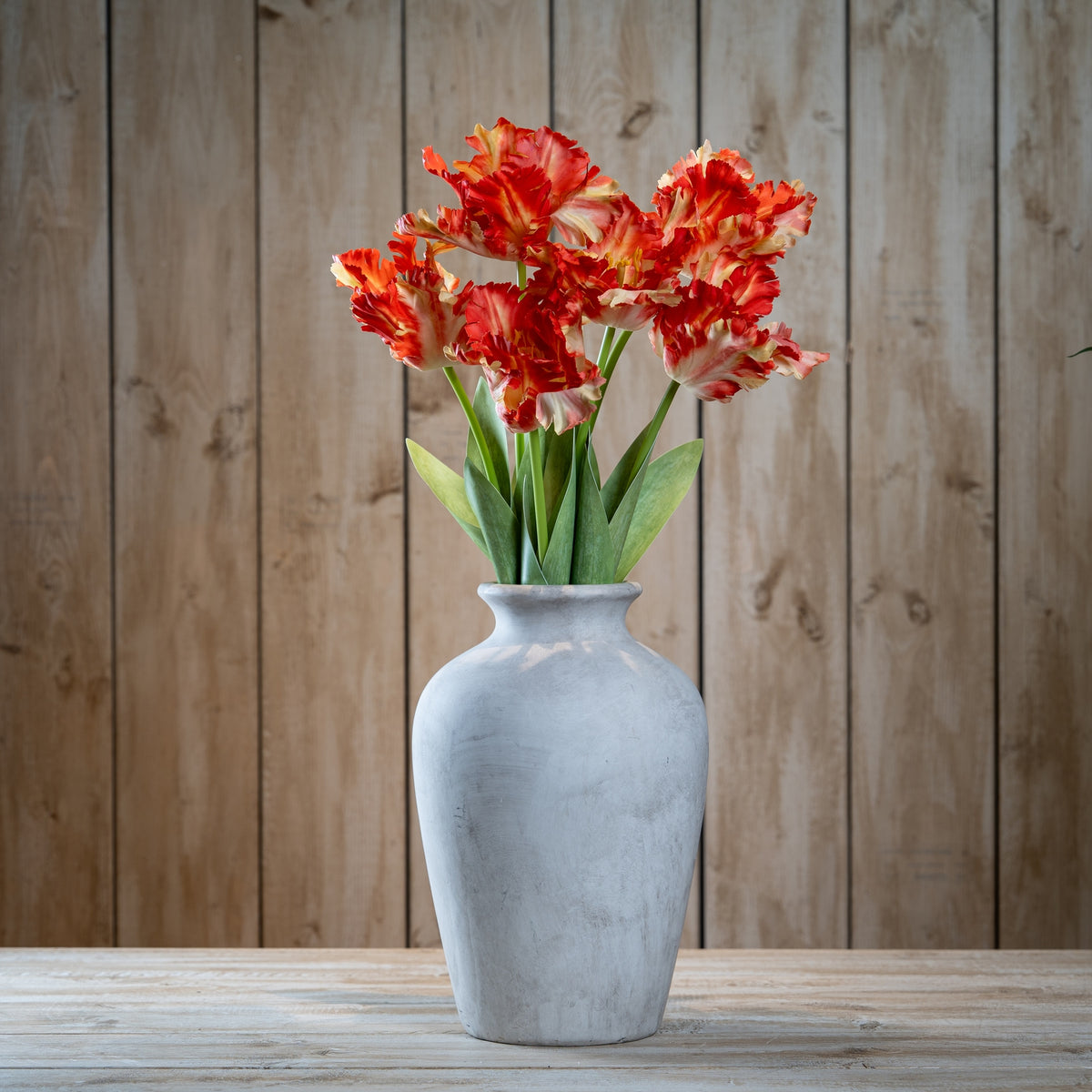 Several orange-red and cream artificial flower stems arranged in a pale grey vase on a wooden table against vertical timber boards.