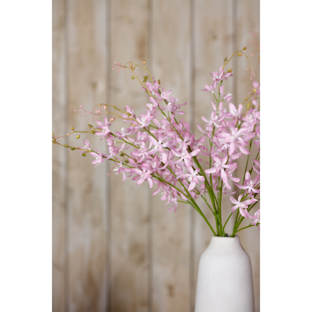 Pale lilac flowering branches spread above a tall white vase in front of light wooden wall panels.