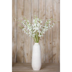 White artificial flower arrangement in a tall white vase on a wooden surface, with vertical wood panelling behind.