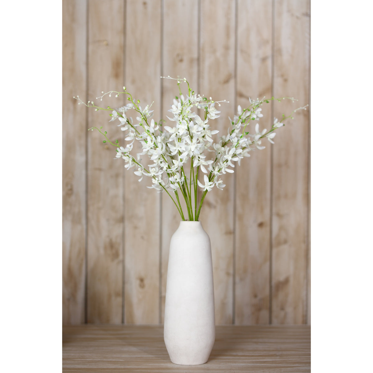 White artificial flower arrangement in a tall white vase on a wooden surface, with vertical wood panelling behind.