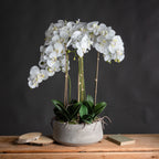 White artificial flower arrangement in a grey oval pot on a wooden surface against a dark wall, with wood slices to the left and a book to the right.
