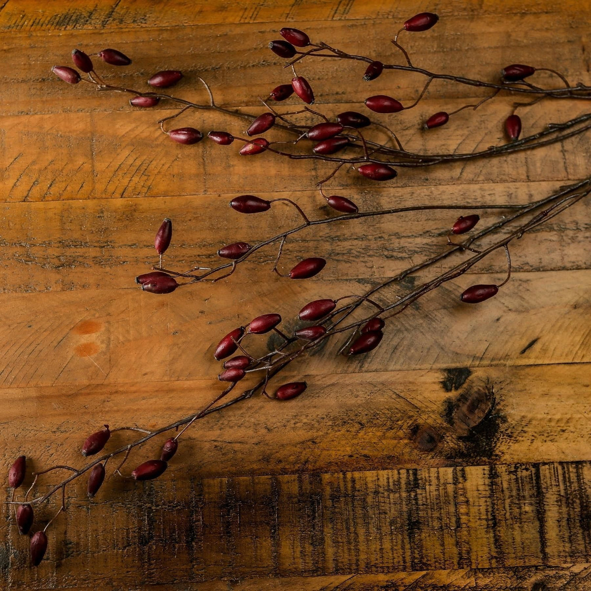 Overhead view of artificial rosehip branches spread diagonally across a textured wooden surface, showing their elongated red forms.
