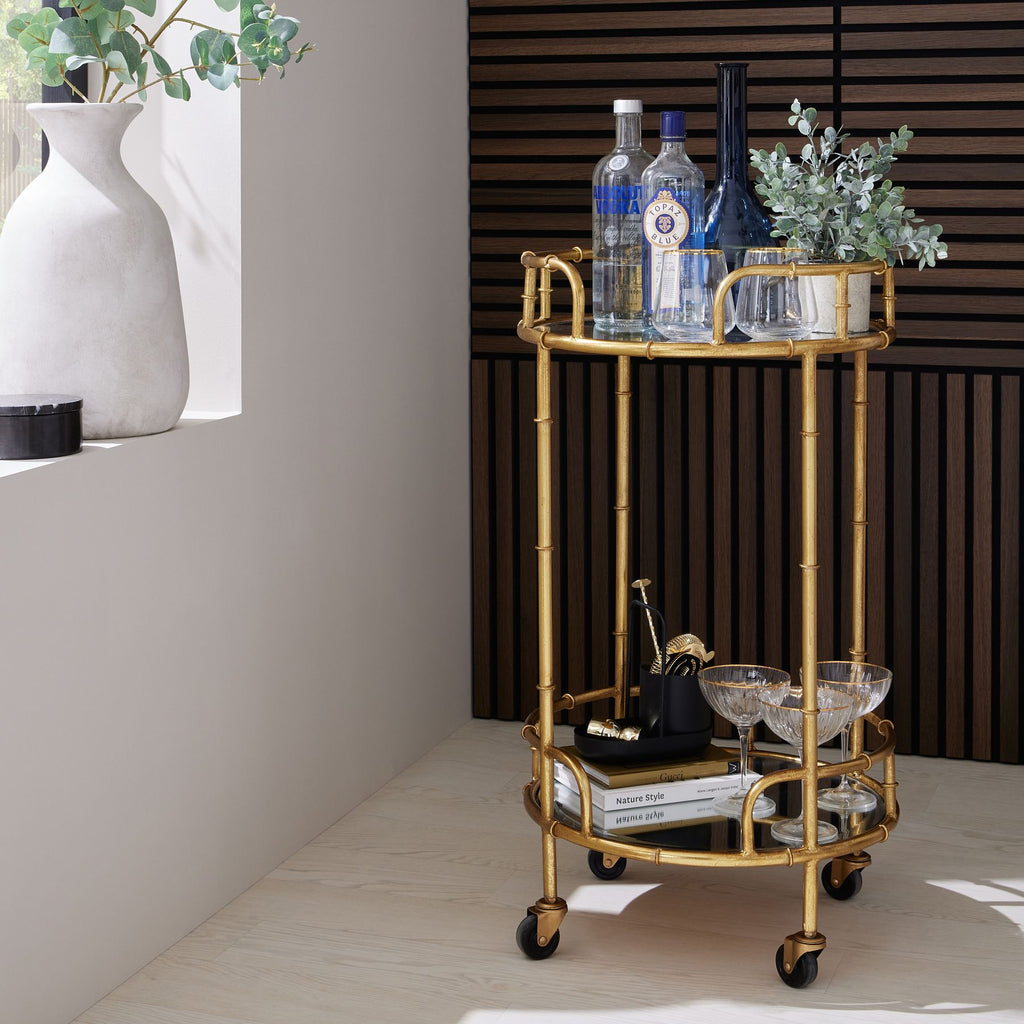 Potted green foliage styled beside bottles and glasses on the upper shelf of a gold-coloured drinks trolley against dark wood slats.