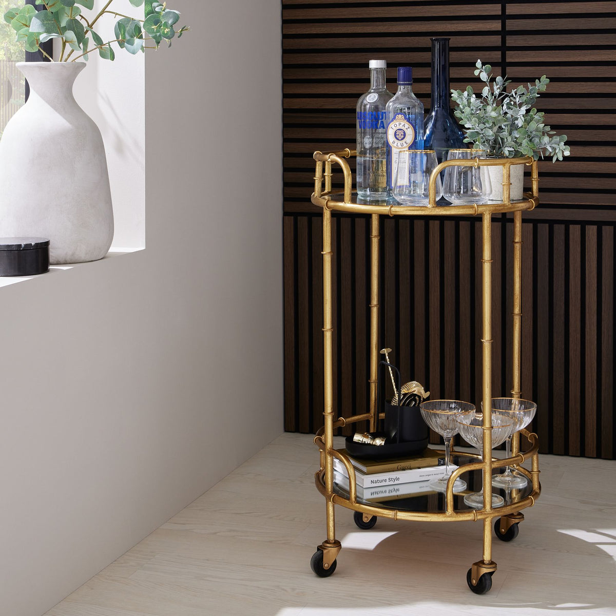 Potted green foliage styled beside bottles and glasses on the upper shelf of a gold-coloured drinks trolley against dark wood slats.