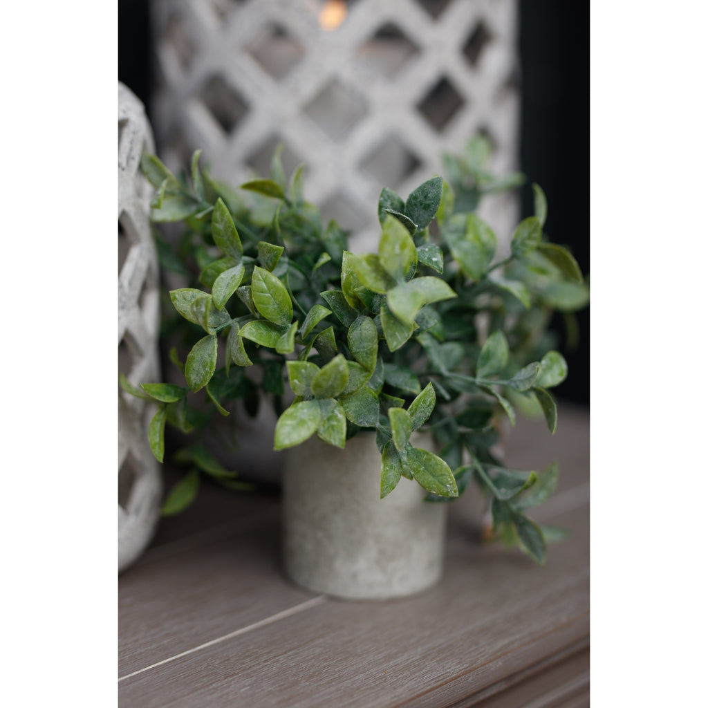 Artificial potted greenery on a wooden surface beside pale decorative objects, with a patterned object behind the leaves.