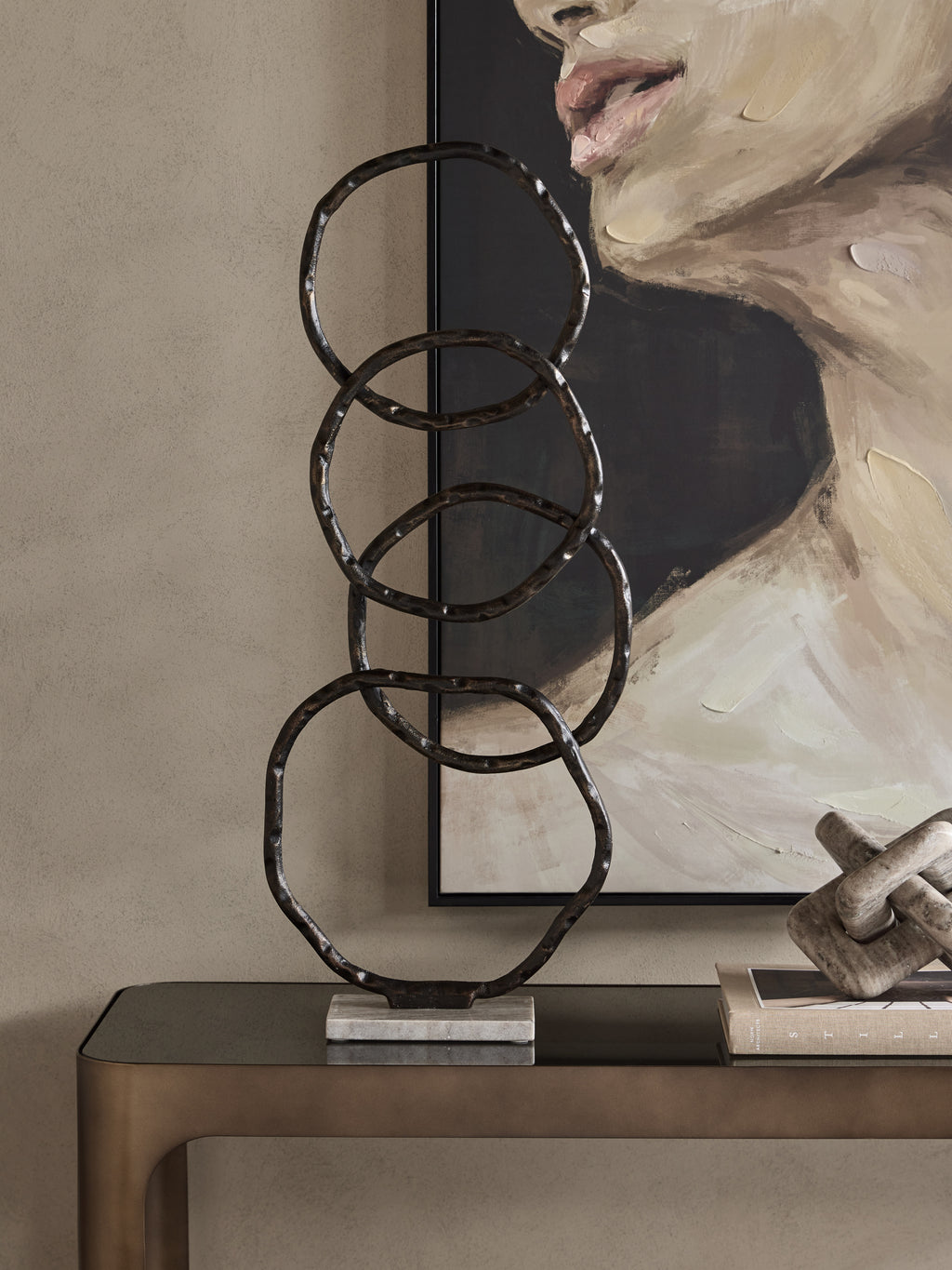 Ring sculpture on a dark console in front of a framed portrait, beside books and a pale decorative knot.