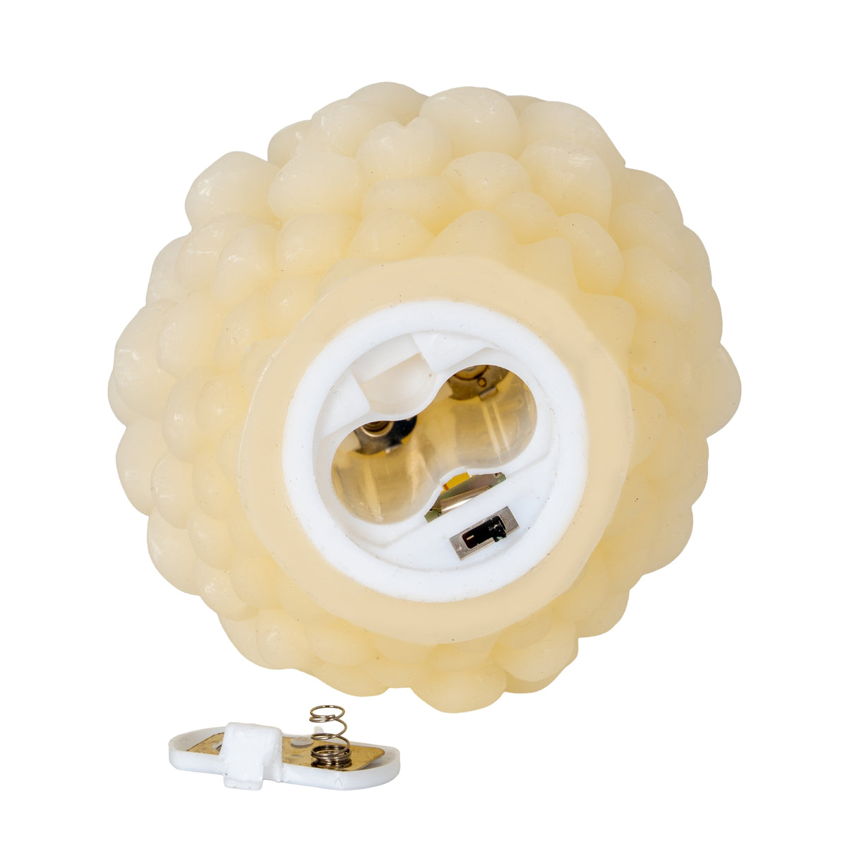 Underside of the cream pinecone LED candle showing an open empty battery compartment and a slide switch, with the removed door and spring contact below.