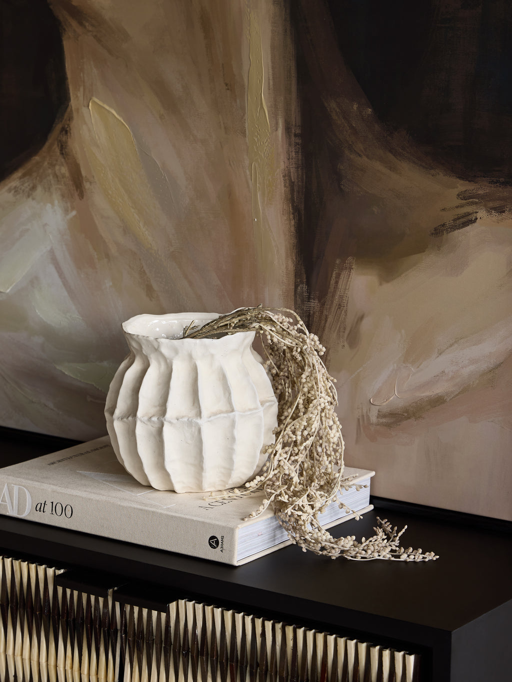 Rounded white panelled vase with trailing dried stems on an art book, set on a dark cabinet in front of an abstract painting.