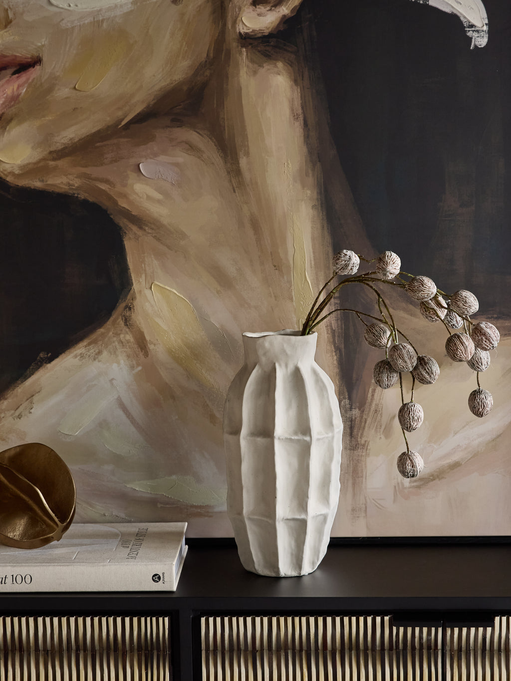 Tall white panelled vase holding arching stems with round pods on a dark cabinet, beside books and a gold-toned object beneath a large portrait.