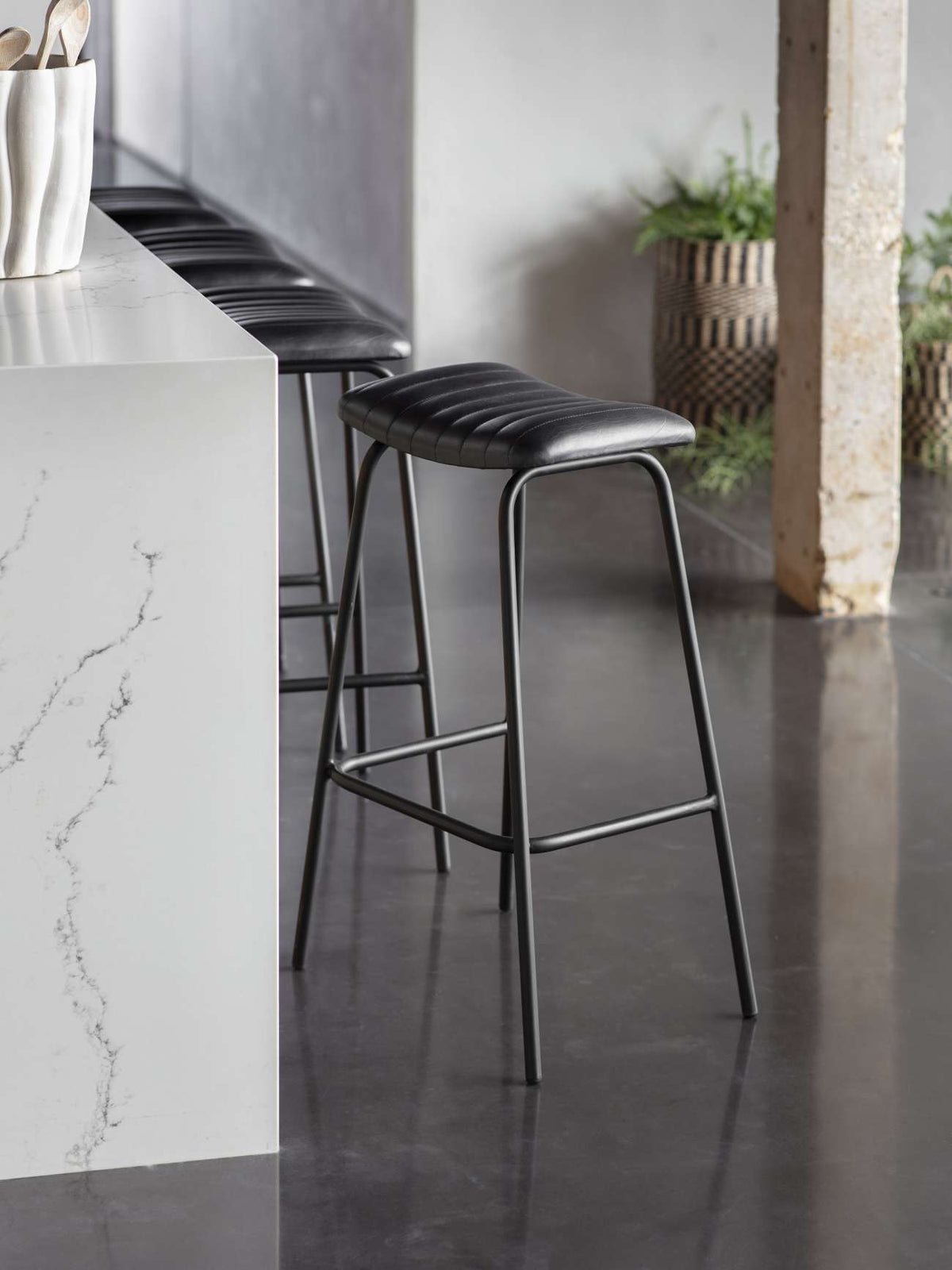 Charcoal stitched bar stools lined along a white veined kitchen island, with dark flooring and woven plant baskets nearby.