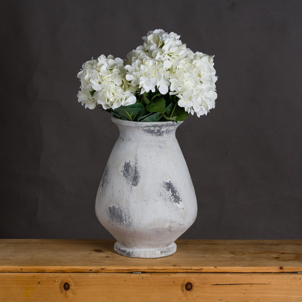Patch Urn Vase styled with white flower heads and green leaves on a wooden surface against a dark grey wall.