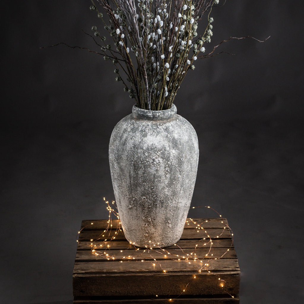Patchneck Vase filled with tall decorative branches on a wooden crate, with string lights around the base against a dark backdrop.